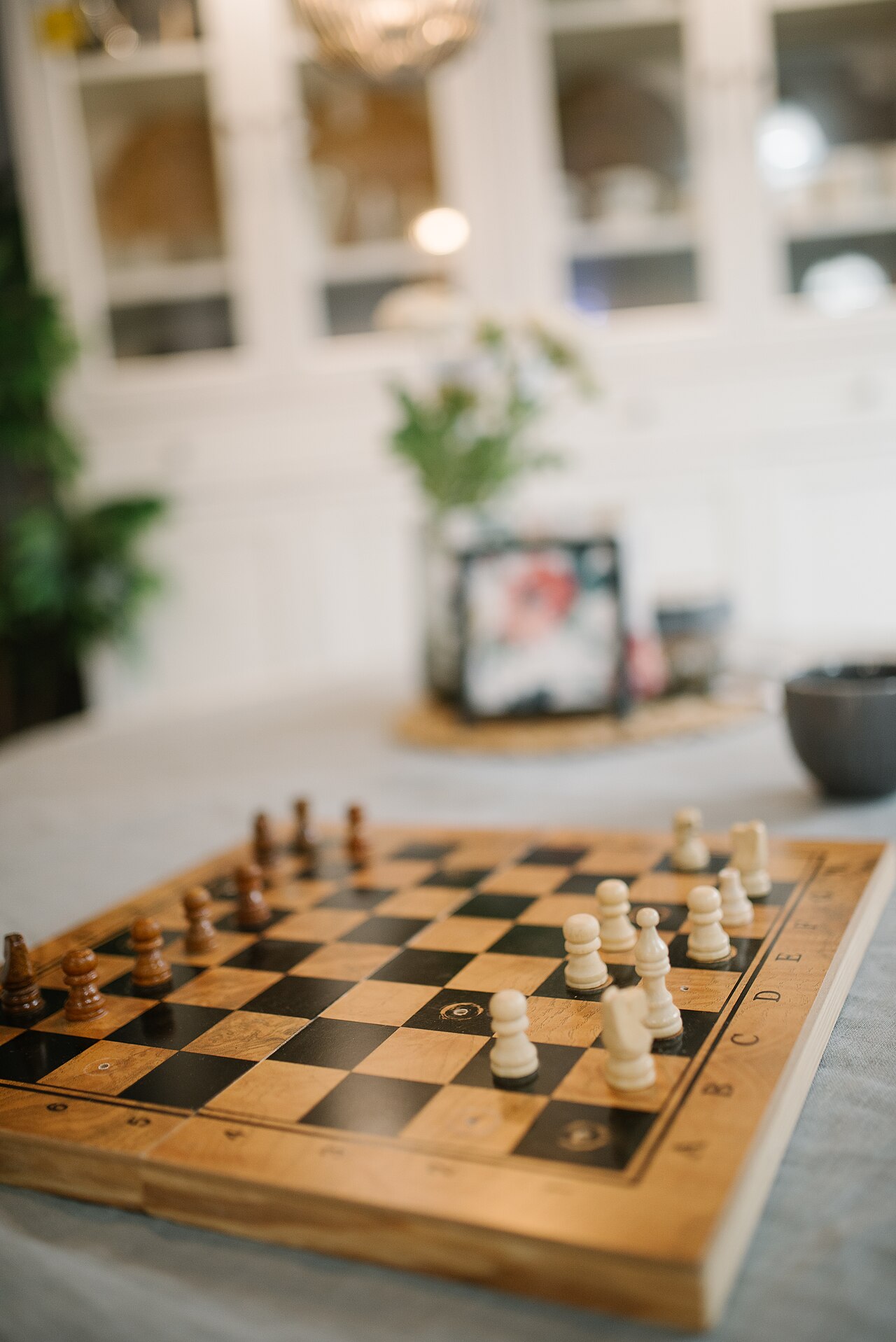 A wooden chess board mid-game on a kitchen table, pieces developed on both sides, in a bright and homely room.